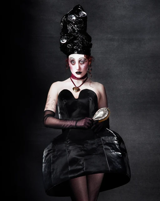 Portrait of a person with exaggerated makeup, wearing a black dress and elaborate black headpiece with reflective accents, holding a hairbrush.
