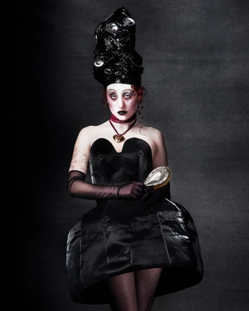 Portrait of a person with exaggerated makeup, wearing a black dress and elaborate black headpiece with reflective accents, holding a hairbrush.