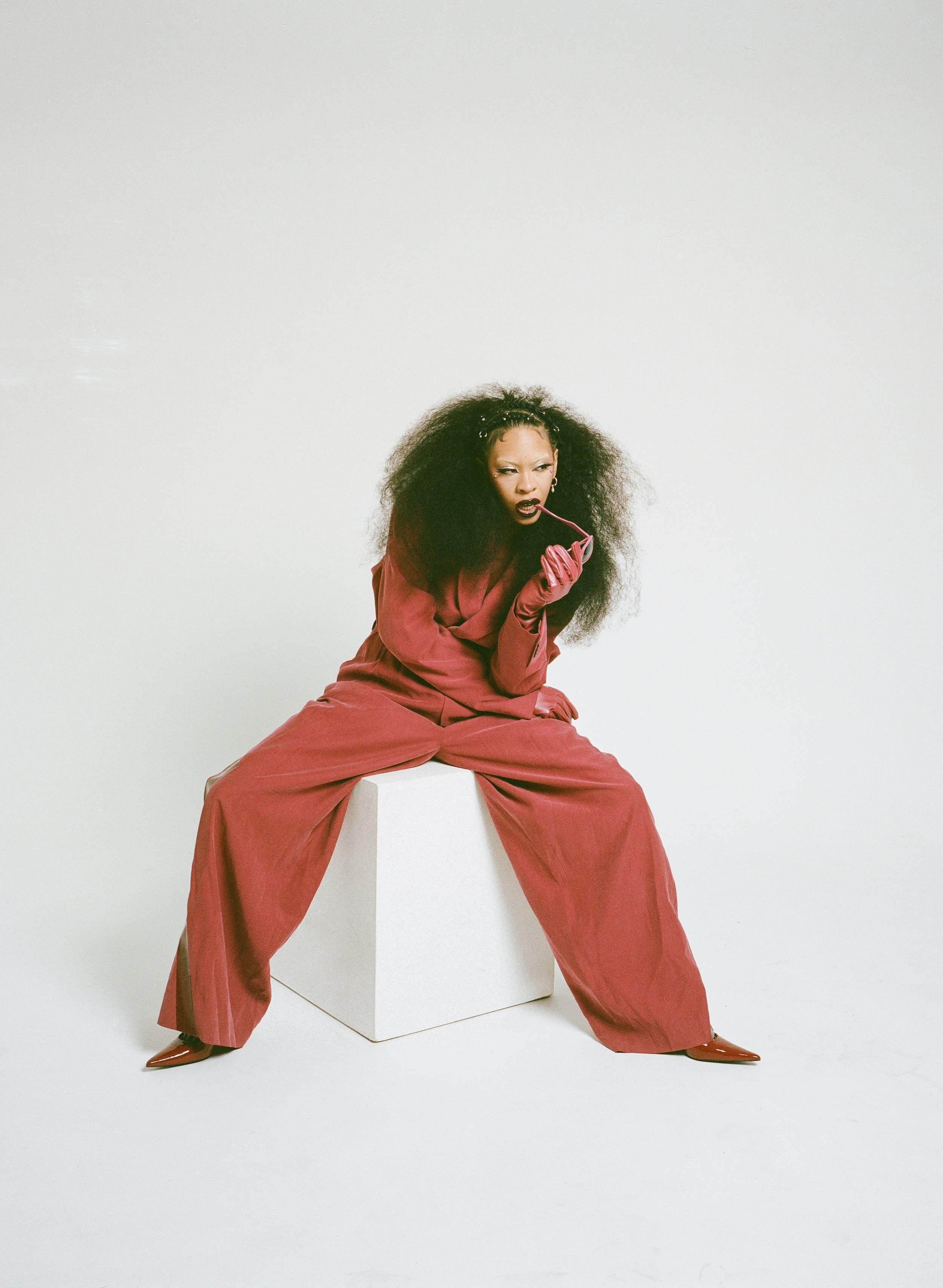 Woman in red pantsuit with large afro sits on white cube, holding glasses in studio. Fashion shot with plain background.