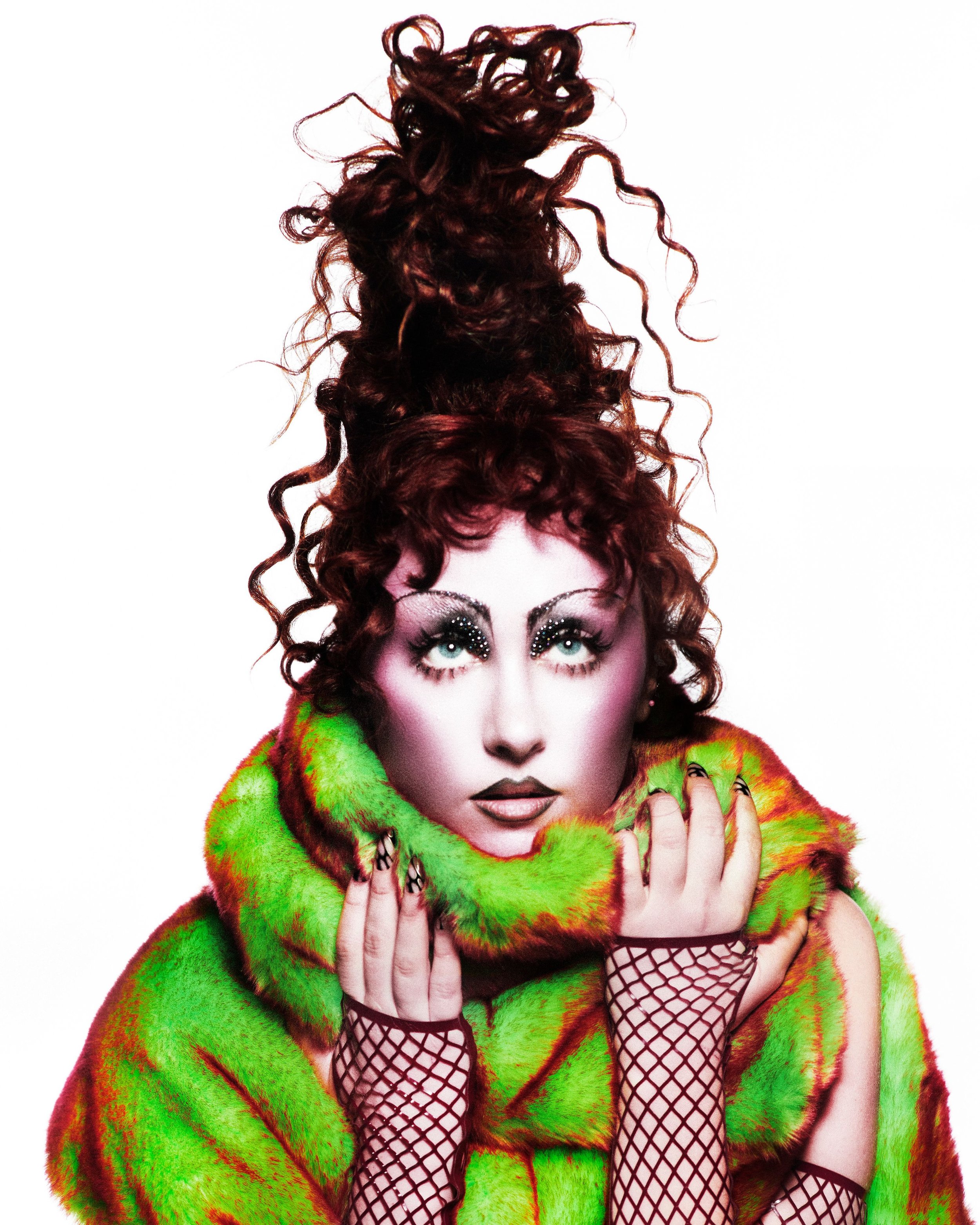 Fashion shot of a woman with an elaborate updo, dramatic makeup, a green and brown fur coat, and fishnet gloves, posing against a white backdrop.