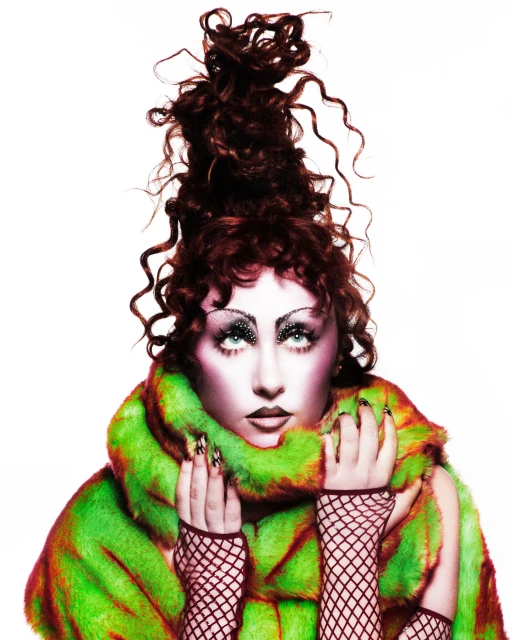 Fashion shot of a woman with an elaborate updo, dramatic makeup, a green and brown fur coat, and fishnet gloves, posing against a white backdrop.