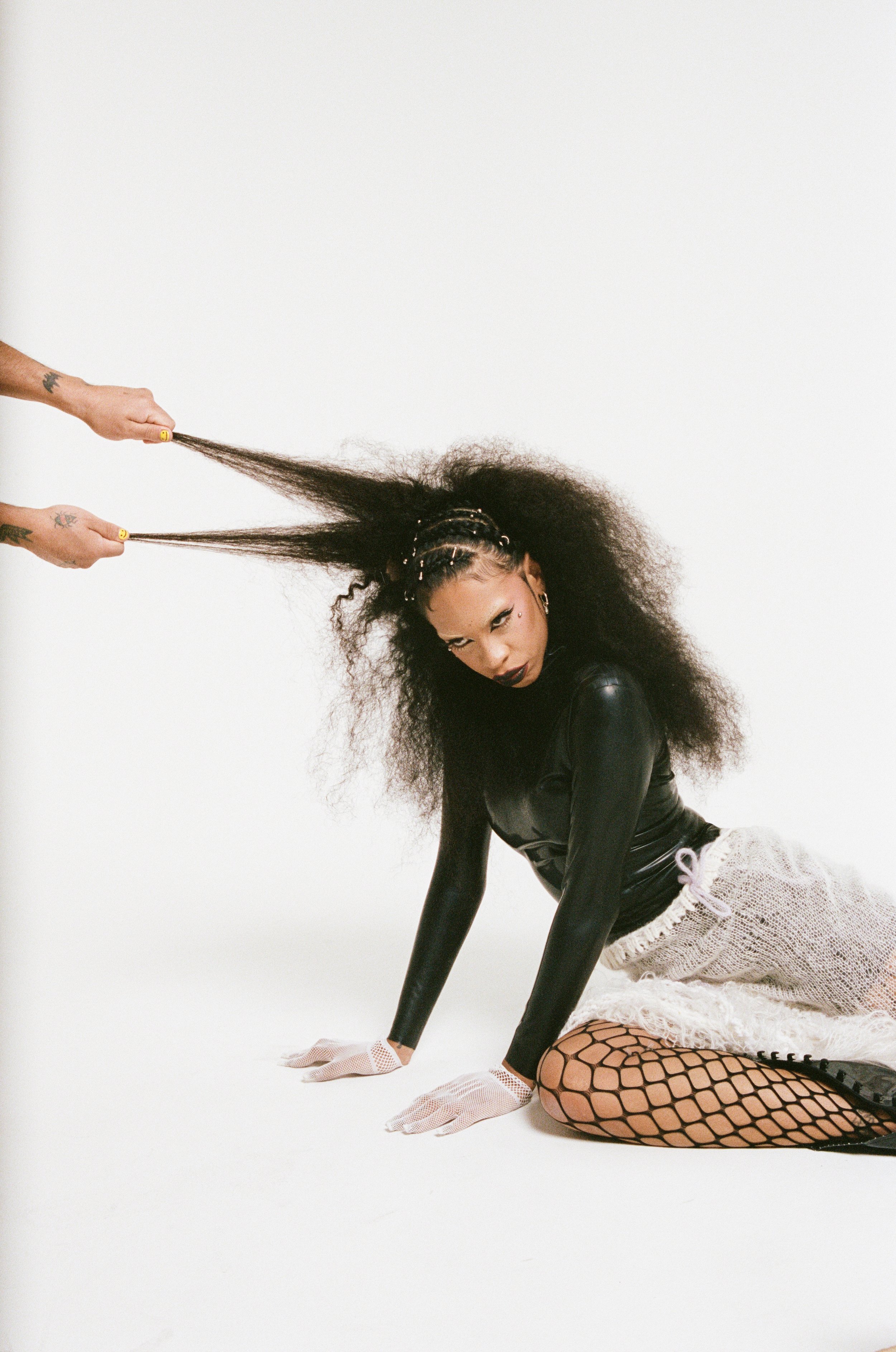 Woman in glossy bodysuit and fishnet tights poses on a white background while hands pull her afro hair out to each side.