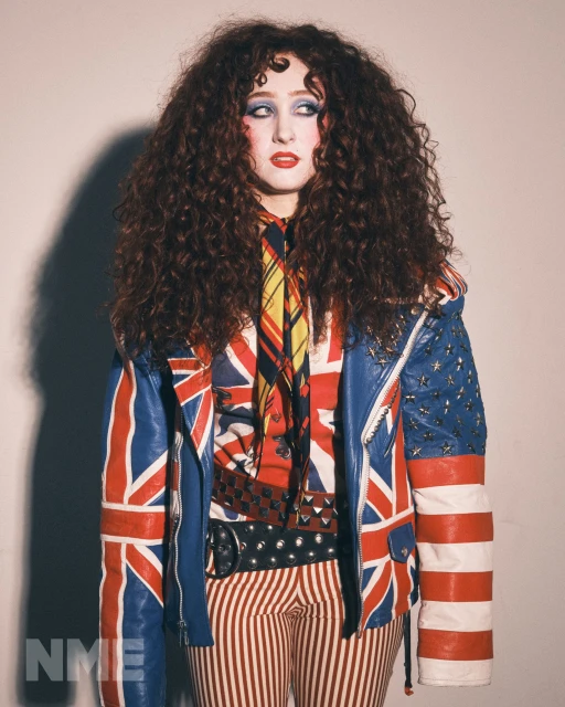 Portrait of a woman with curly hair, wearing a Union Jack and Stars & Stripes jacket and striped pants, against a plain background.