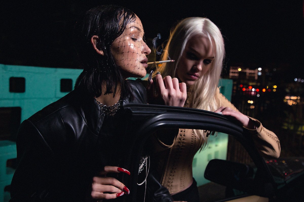 Two women pose in front of a car at night; one smokes. The other rests her arm on the vehicle; city lights glow in the background.
