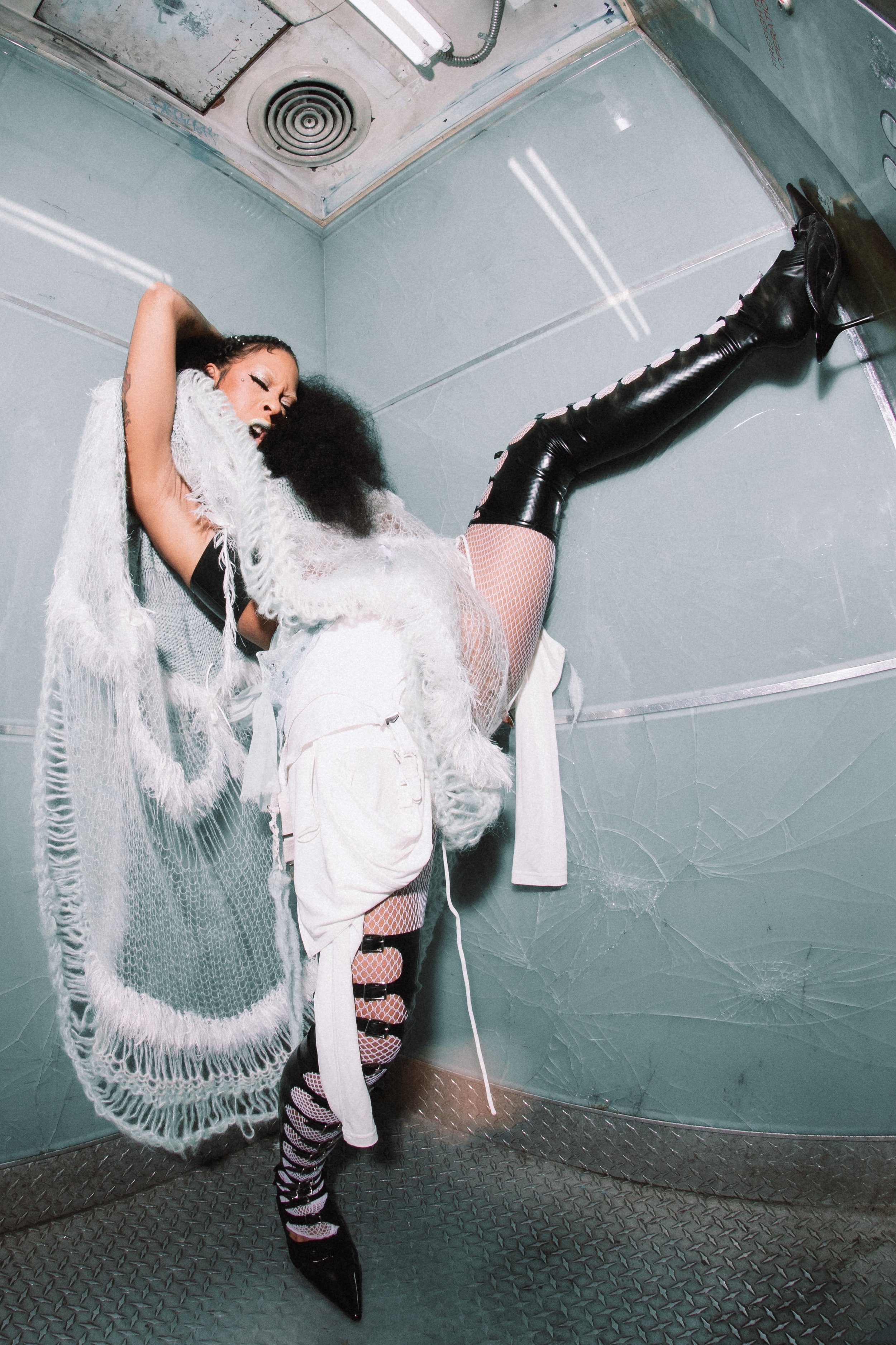 Woman in an elaborate white outfit and black, thigh-high boots poses against a blue-grey wall, one leg raised in a dramatic fashion shoot.