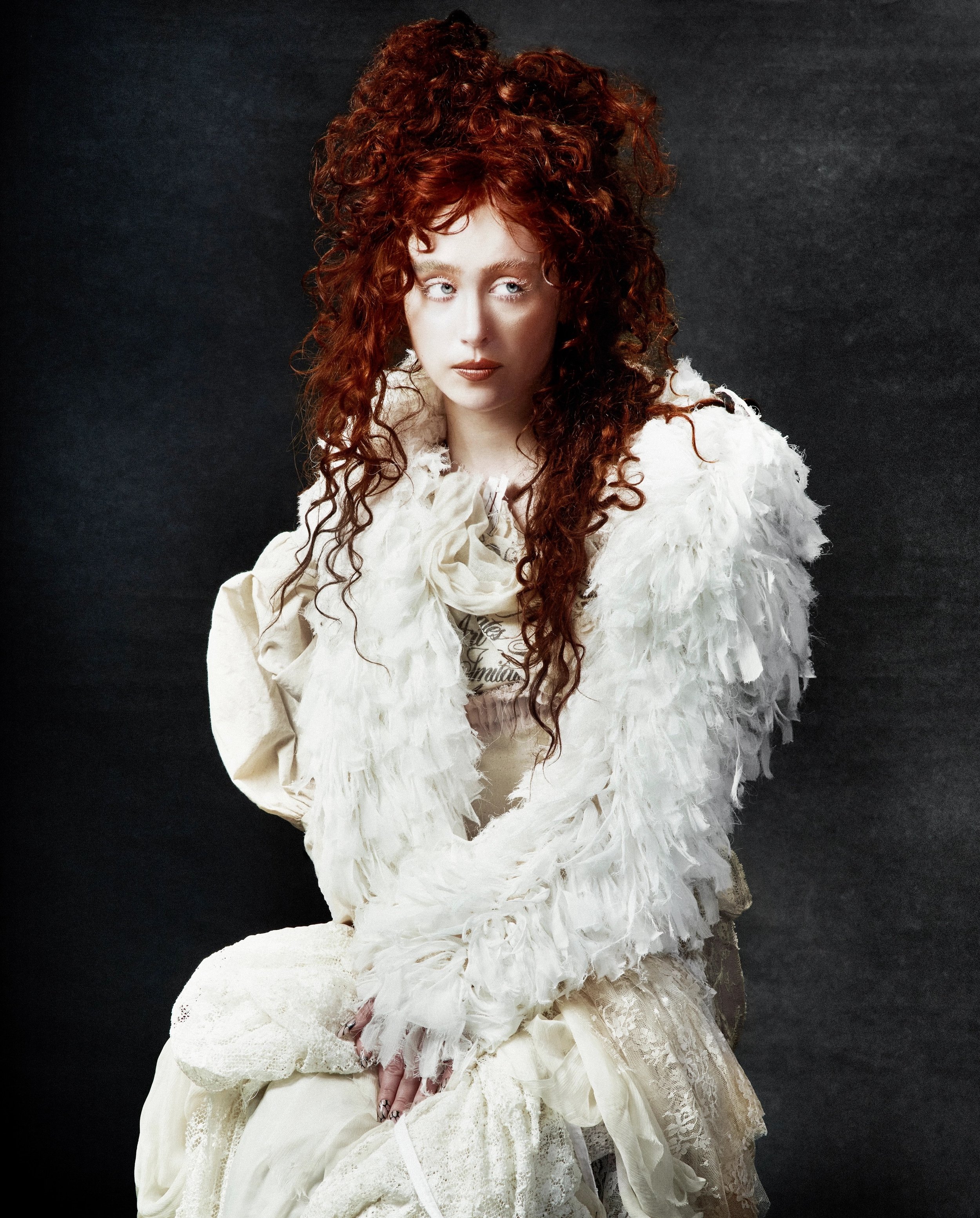 Portrait of an adult woman with red curly hair wearing a feathered shawl and lace gown in a dimly lit, dark grey environment.