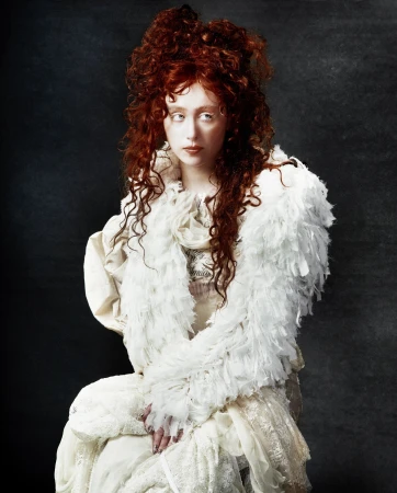 Portrait of an adult woman with red curly hair wearing a feathered shawl and lace gown in a dimly lit, dark grey environment.