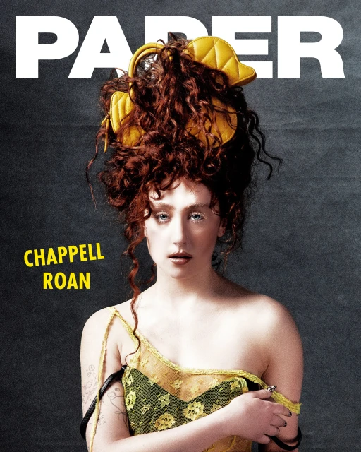 Editorial-style shot of Chappell Roan on PAPER Magazine cover with statement red curly hair and yellow hat-like accessory. 