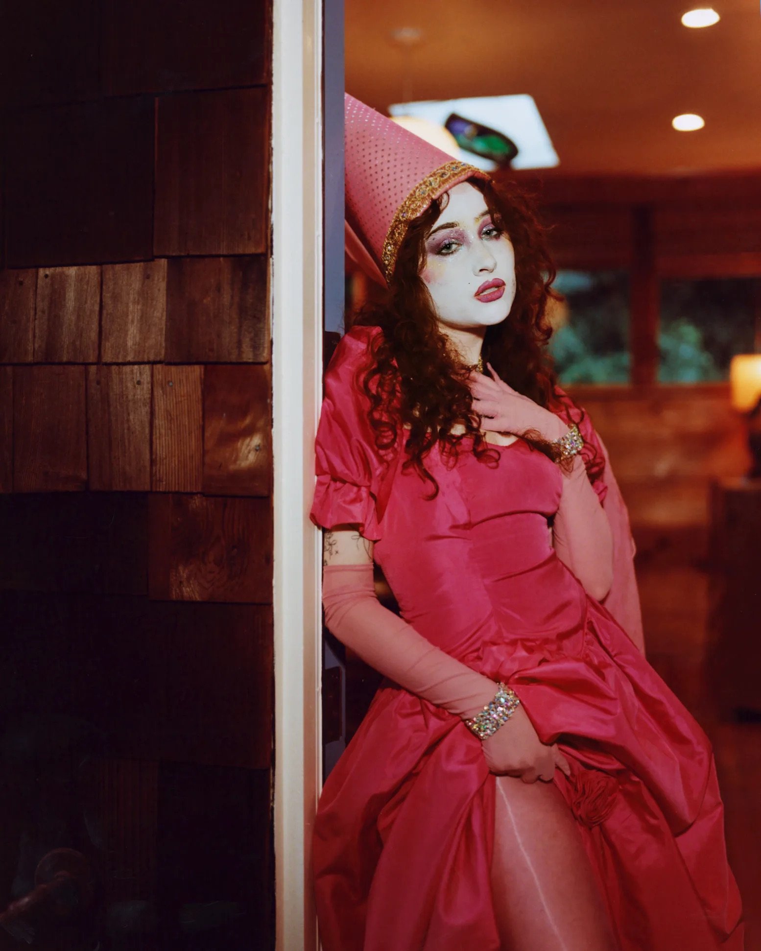 Woman in a pink dress and cone hat leans against a wall, showing her leg with white makeup and red lipstick, looking at the viewer.