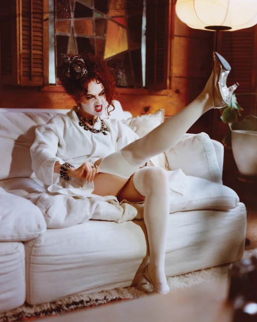 Woman in robe reclining on sofa, silver high heel, white thigh-high stocking, vintage style, ornate jewelry, with a neutral indoor setting.