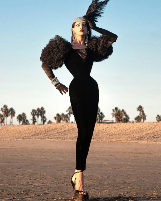 A slender woman in a black dress and platform heels poses on a beach with palm trees in the background under a clear sky.