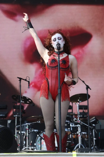 A person sings on stage in a red costume. Red boots and fishnet stockings are worn. A microphone and background equipment are visible.