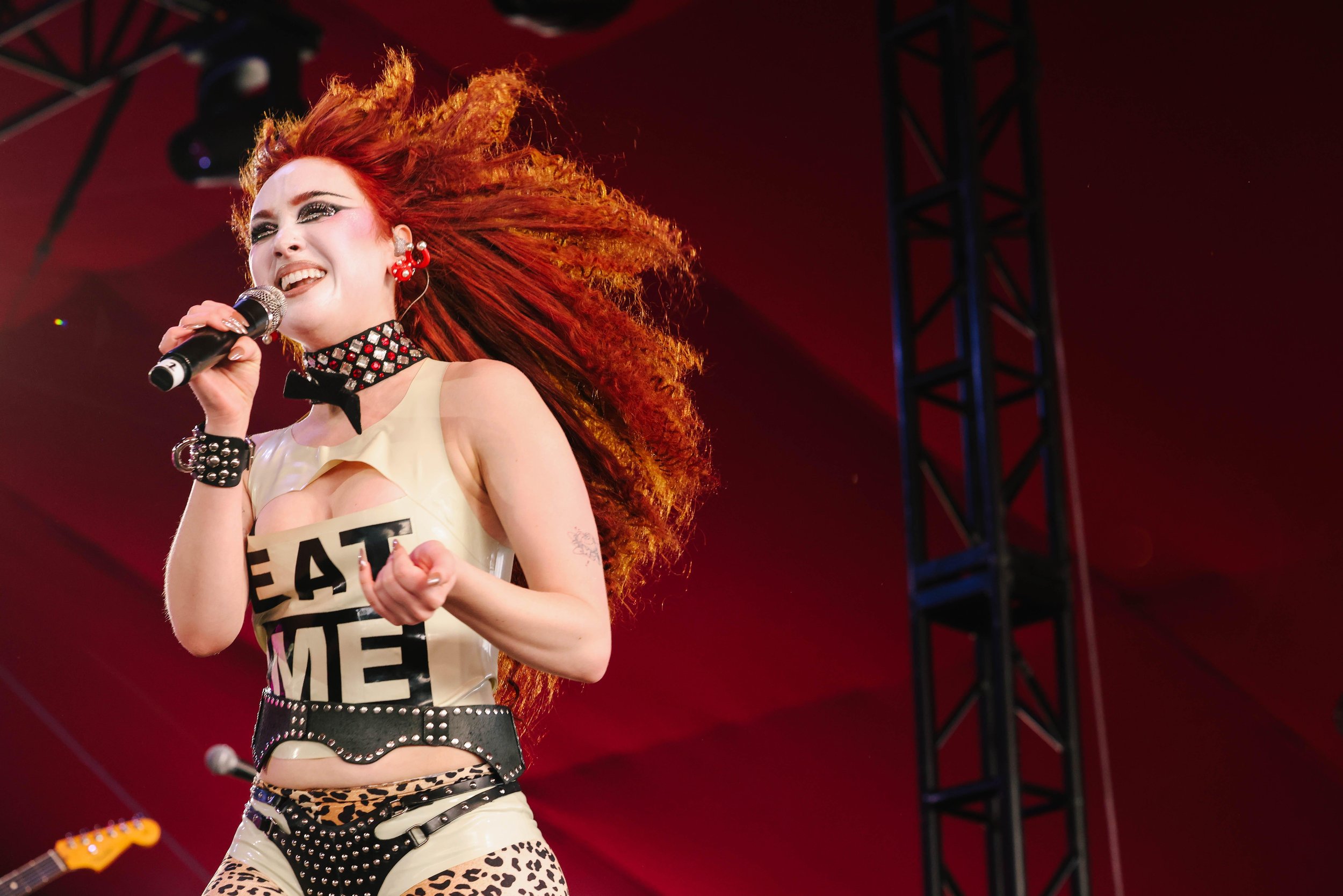 Female singer with red hair performs on stage with a microphone wearing a white top, patterned bottoms, and a studded belt and choker.