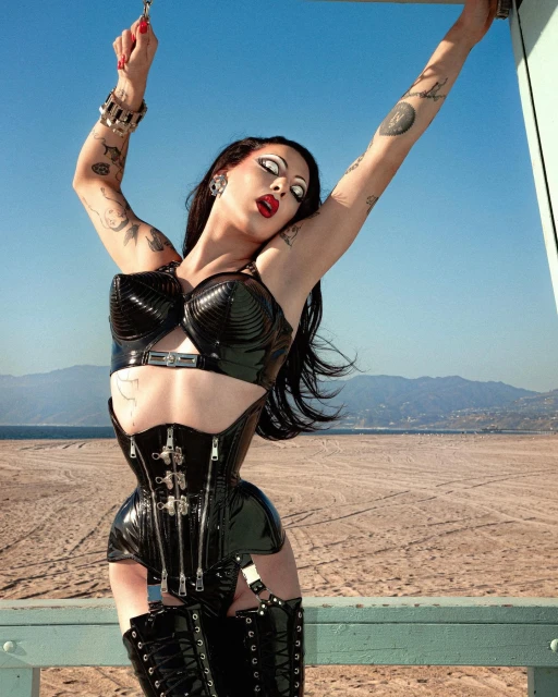 Woman in a black latex corset and thigh-high boots poses on a beach pier with a blue sky, sandy beach, and mountains in the background.