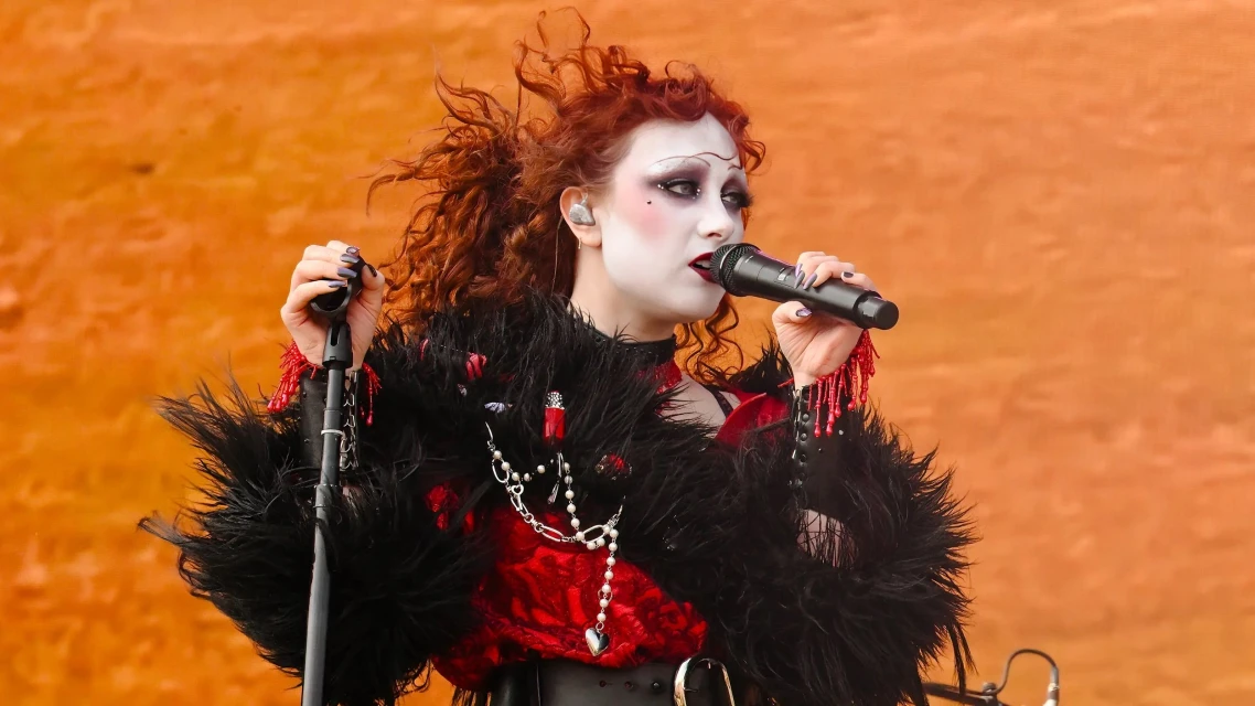 A woman with curly red hair and white makeup, wearing a black and red outfit, sings into a microphone against an orange background.