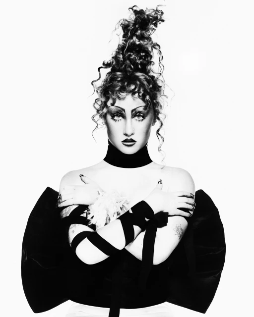 Striking black and white studio portrait of a woman with elaborate curly updo, theatrical makeup, and a dramatic ribbon-wrapped garment.