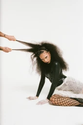 Model with voluminous dark hair being pulled by hands, wearing black top and textured skirt against a plain white background