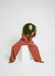 Model with voluminous dark curly hair sitting on a white cube, wearing a matching red pant suit and holding sunglasses against a white background.