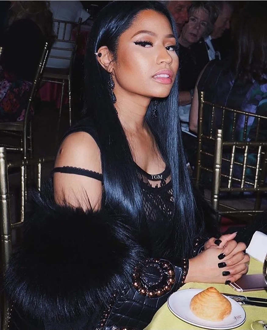 Close-up of Nicki Minaj at a dining table wearing a black lace top and a fur stole, with a dessert plate in the foreground.