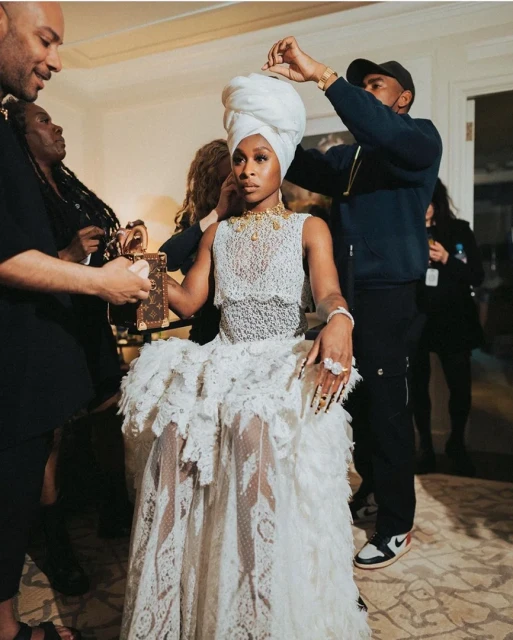 An adult female in an elegant white lace dress and turban is being attended to by stylists in an indoor setting, preparing for an event.