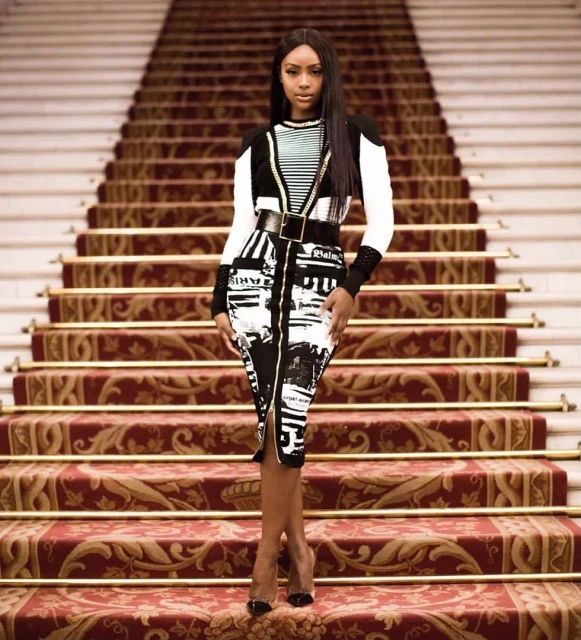 A woman in a black and white patterned dress stands on a red and gold staircase, posing and looking at the camera.