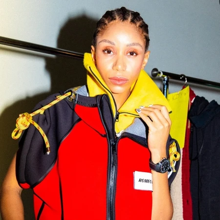 Model with braided hair wearing a red, black, and yellow hooded jacket with patterned nails.