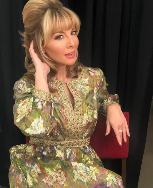 Blonde woman in patterned gown with floral detail sits indoors near dark backdrop, smiling, and resting her hand on her hair.