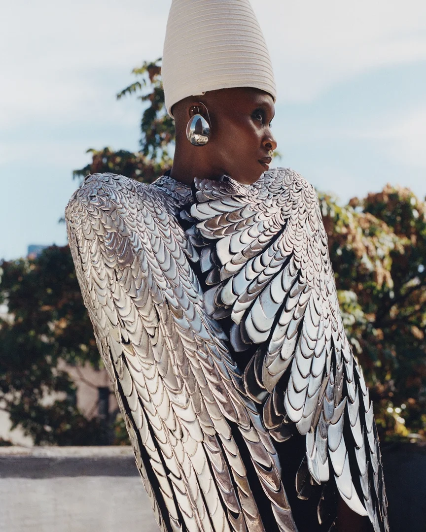 An adult female with shaved head poses in a silver feather-like dress with a large white hat against a blurry background of trees and buildings.