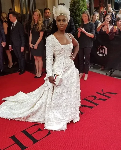 A woman in a white gown poses on a red carpet outside a building, with onlookers and photographers in the background.