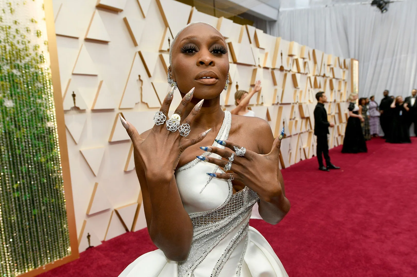 Cynthia Erivo poses on a red carpet wearing a white gown and large rings. Several other individuals are in the background.