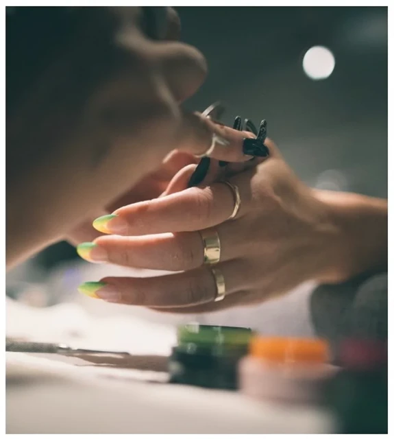 Close-up of a nail technician applying nail polish to a client with gold rings on her fingers and yellow and green tips on her nails.