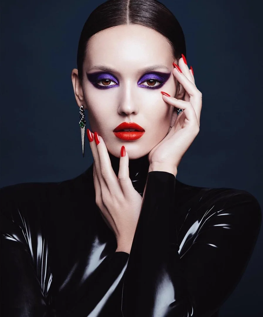Fashion portrait of a woman with red lipstick and striking purple eye makeup, adorned with long green earrings, posing indoors against a dark backdrop.