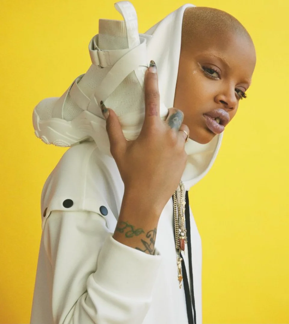 Woman poses in a white hooded jacket with an all-white high-top sneaker resting on her head against a bright yellow backdrop.