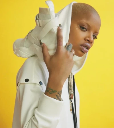 Woman poses in a white hooded jacket with an all-white high-top sneaker resting on her head against a bright yellow backdrop.