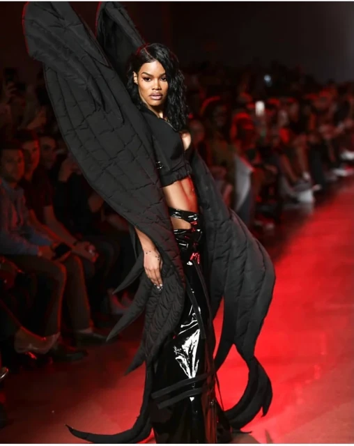 Runway fashion show featuring a Black female model wearing a black crop top, skirt, and large wings, walking in a crowded venue.