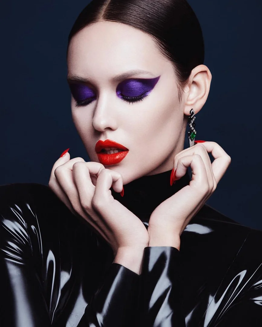Woman with dramatic makeup and glossy black top in studio. Purple eyeshadow, red lips, red nails, and statement earrings.