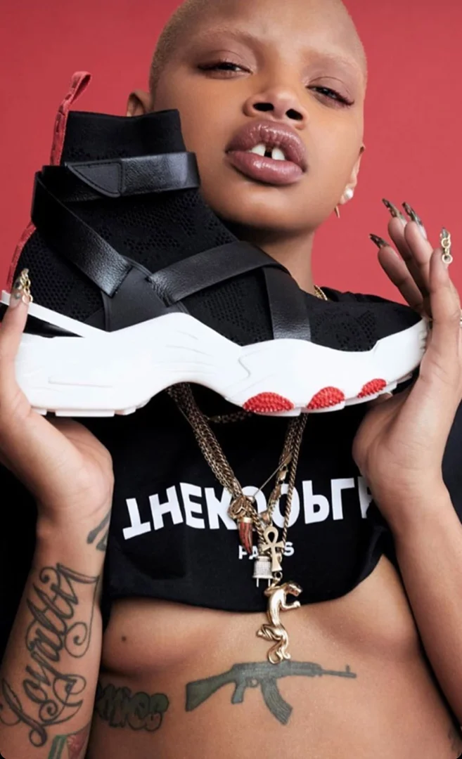 Woman holding black and white high-top sneaker against a red backdrop. She wears gold jewelry and a black crop top with tattoos.