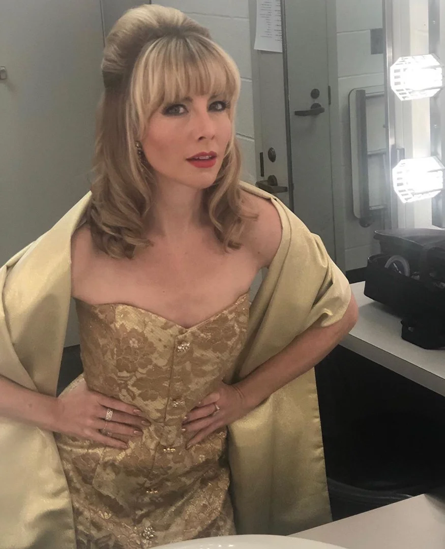 Woman in an ornate gold dress and shawl poses in a dressing room with Hollywood-style lighting, looking at the camera.
