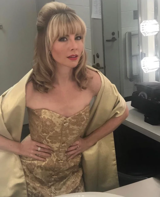 Woman in an ornate gold dress and shawl poses in a dressing room with Hollywood-style lighting, looking at the camera.