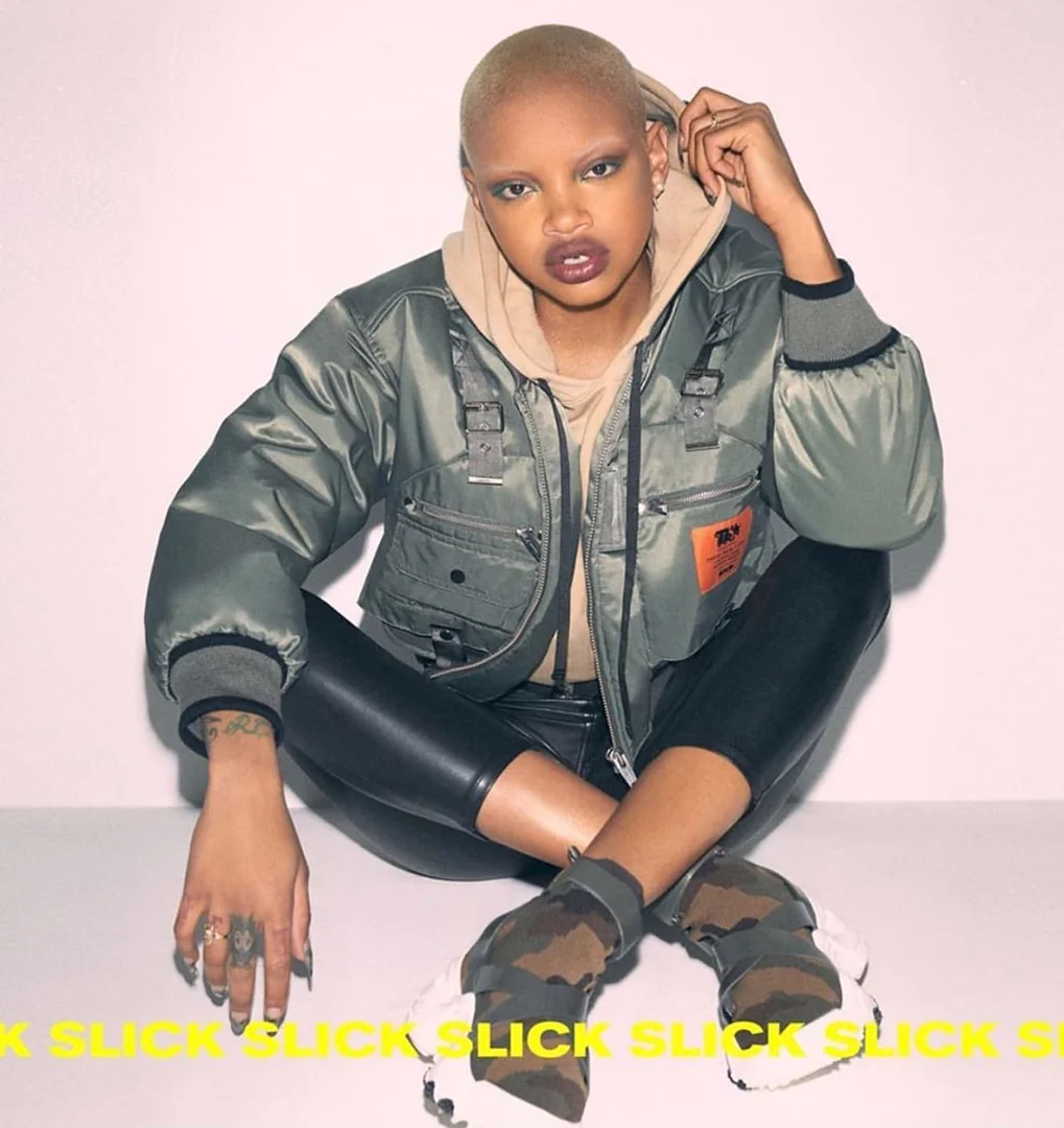Blonde woman sits in olive drab jacket, tan hoodie, black pants, and camo shoes against white background. Fashion shot.