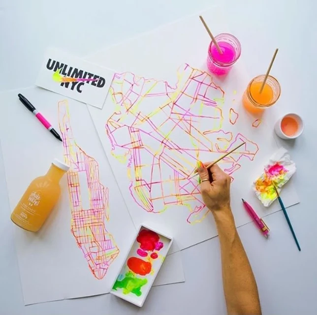 Overhead shot of person painting maps of New York City boroughs with paintbrushes and vibrant colors on a white surface with art supplies.