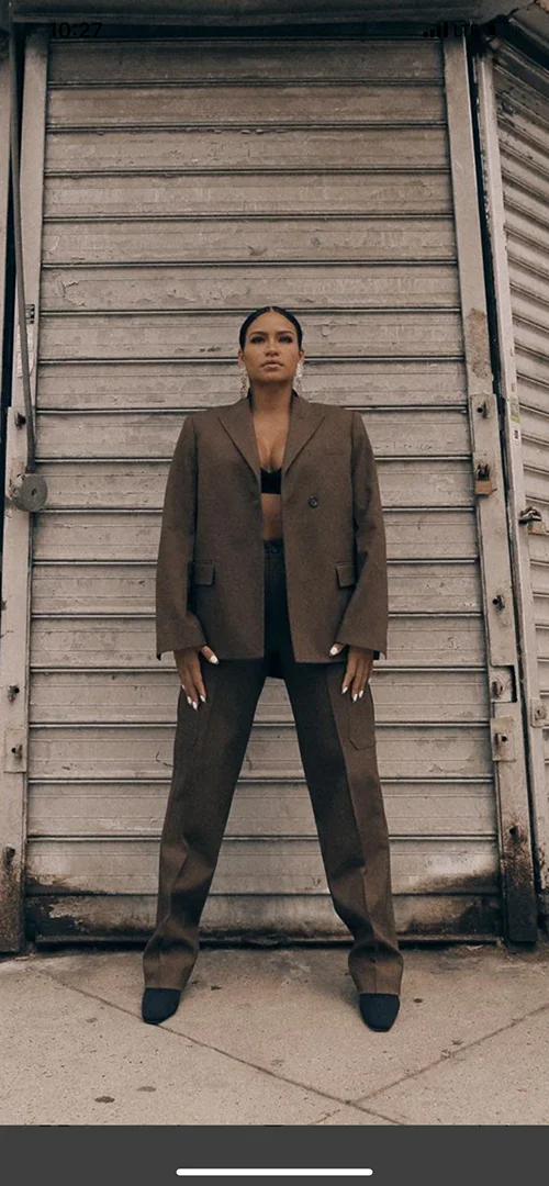 Adult female in a brown suit with an open jacket stands in front of a textured metal wall on an overcast day.