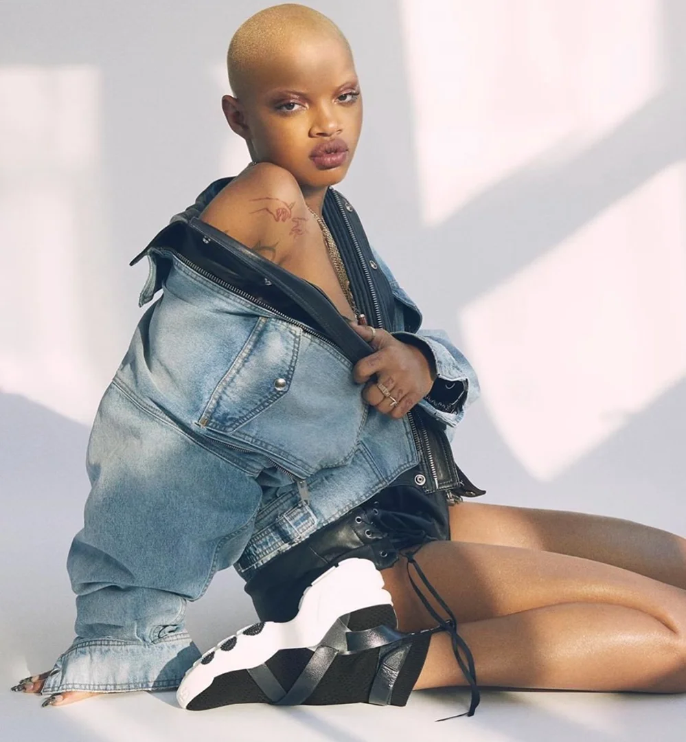 A bald, young adult woman with tattoo on shoulder sitting, wearing a denim jacket and black and white shoes in a photo studio.