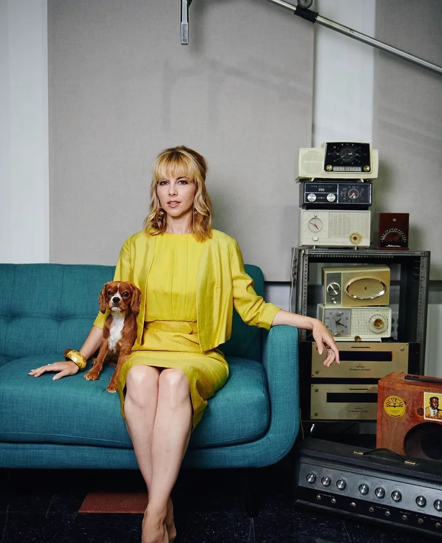 Portrait of an adult woman in a yellow suit with a Cavalier King Charles Spaniel on a turquoise couch surrounded by audio equipment.