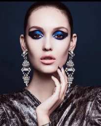 Close-up beauty portrait of a woman wearing dramatic blue eyeshadow and crystal drop earrings with dark metallic nails.