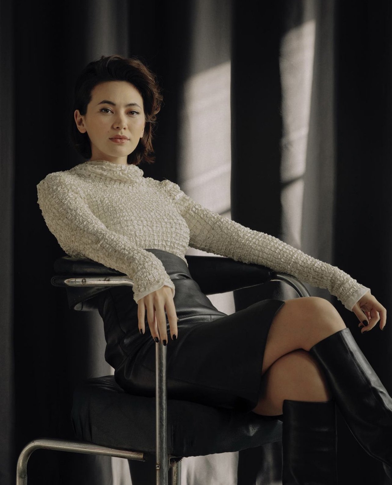 Portrait of a woman with short hair in a white textured shirt, black skirt and black boots, sitting in a chair against a dark backdrop.