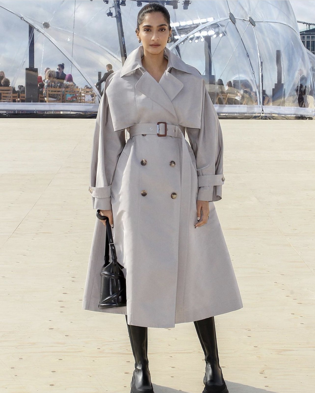 A woman in a light gray trench coat and black boots poses outdoors in front of a translucent structure on a wooden platform.