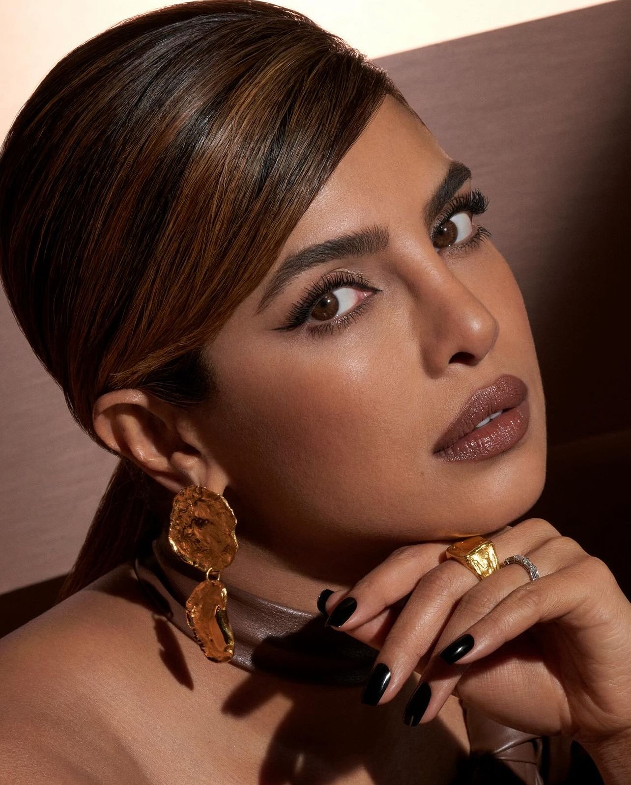 Portrait of adult woman with dark hair, dark lipstick and black nail polish, wearing gold jewelry against a soft brown backdrop.