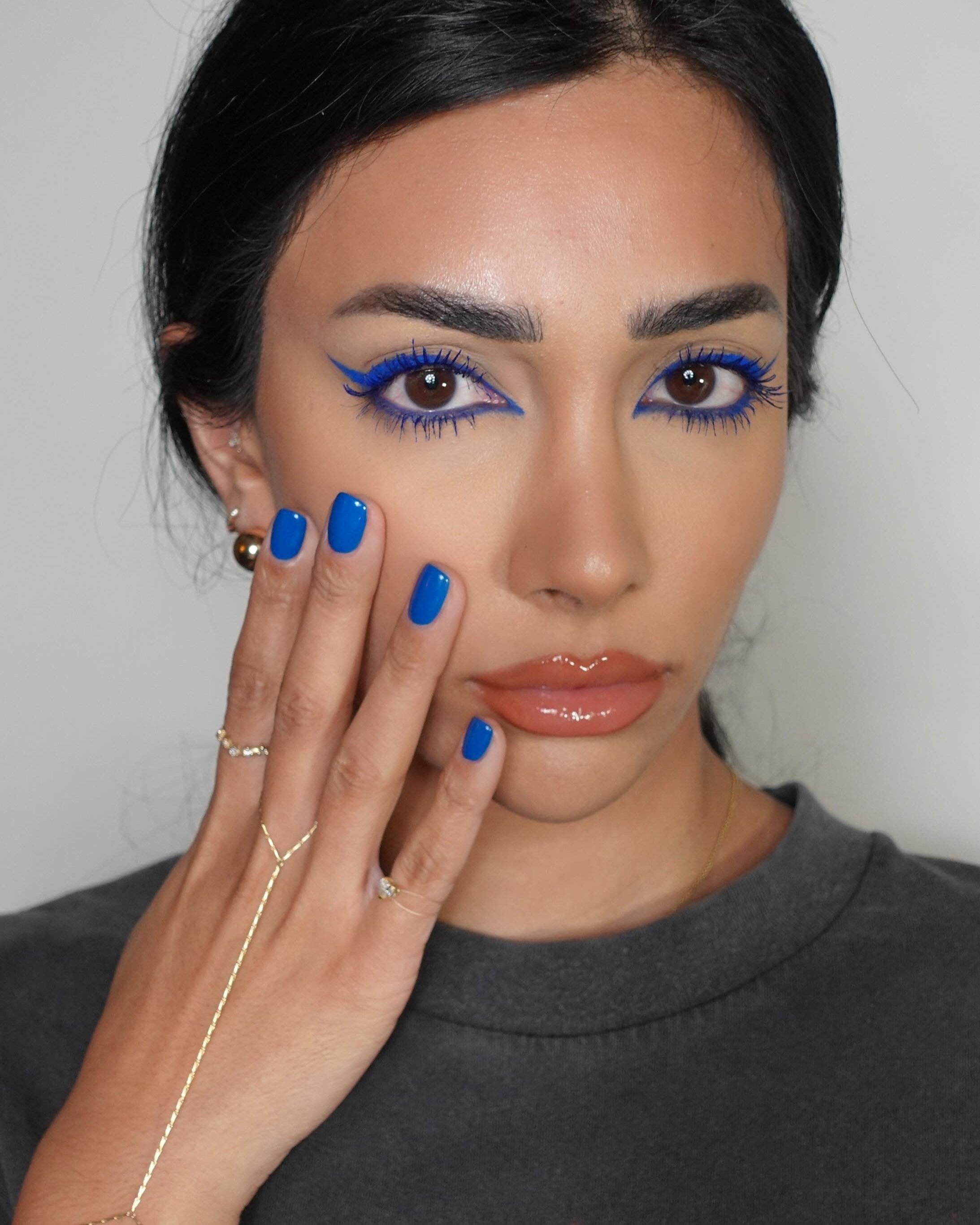 Close-up of a woman with bright blue eyeliner and mascara, matching blue nail polish, and shiny lips, showcasing beauty and makeup.