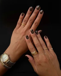 Close-up of hands with glossy dark brown metallic nails wearing gold rings and a gold watch against black